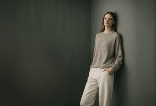 Woman in a taupe knit sweater and cream trousers leaning against a dark grey wall